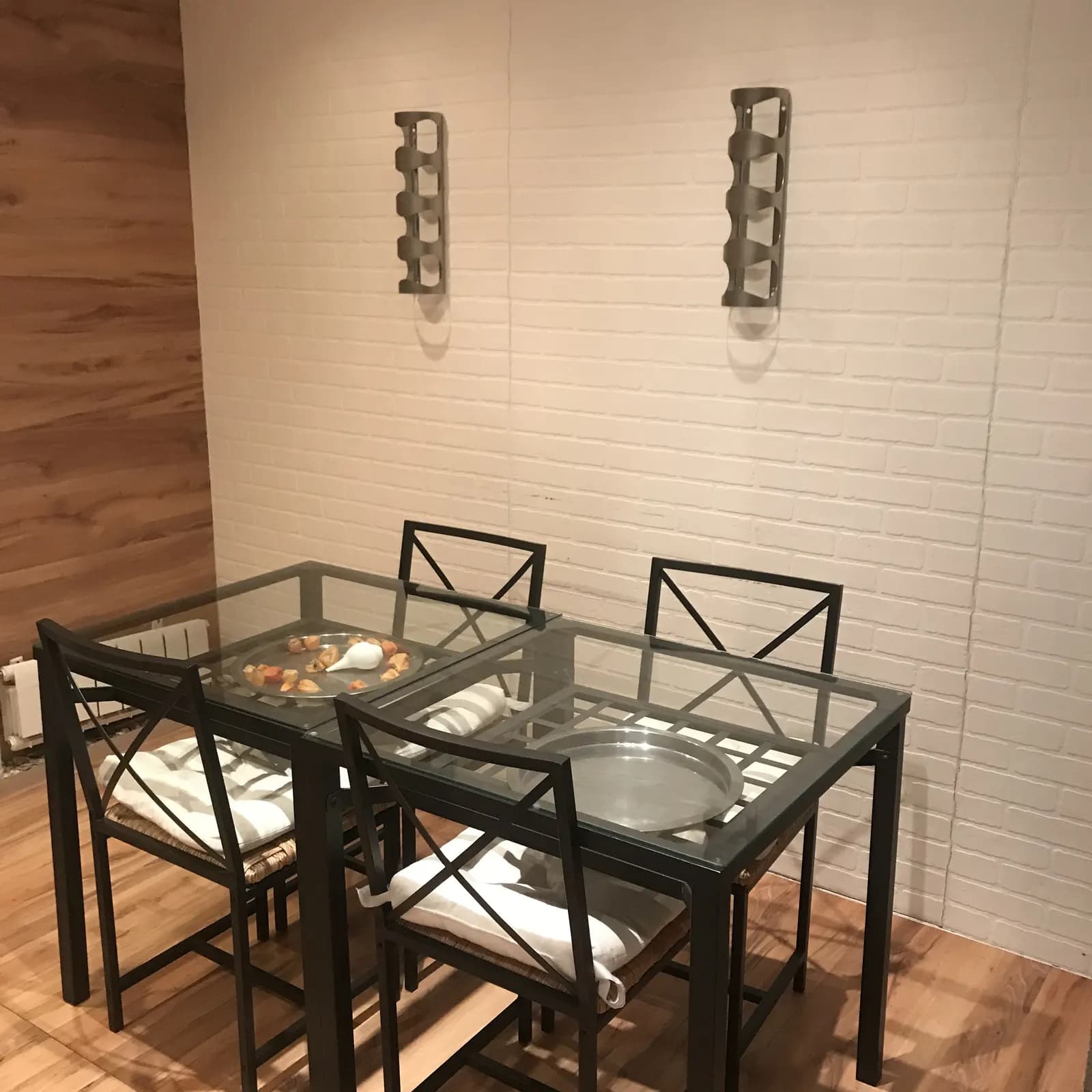Glass dining table with black metal chairs against a white brick wall and wine rack